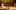 19-Small-Island-2-BrinkhoffMoegenburg-2000x1000-1-aspect-ratio-16-10 On a large stage, a woman in a white dress relaxes on a bench, to the right behind her, another woman in a colourful dress watches on, behind them a line of figures stand at the back of the stage with silhouettes suggesting a crowd projected onto the back wall.