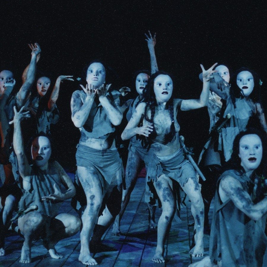 2002_Bacchai_Masks_Manuel Harlan_2000x1000 Performers on stage wear tattered, monochromatic costumes and eerie, white masks, their bodies contorted in expressive, dramatic poses. The lighting is dark, creating a haunting and theatrical atmosphere.