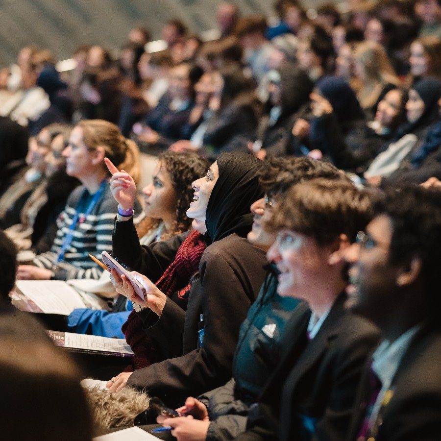 24-StudentConference-pointing-Emma-Hare-2000x1000 A young person pointing to the stage whilst another person smils and looks upwards, they are sitting in the Lyttelton Theatre which is full of other young people