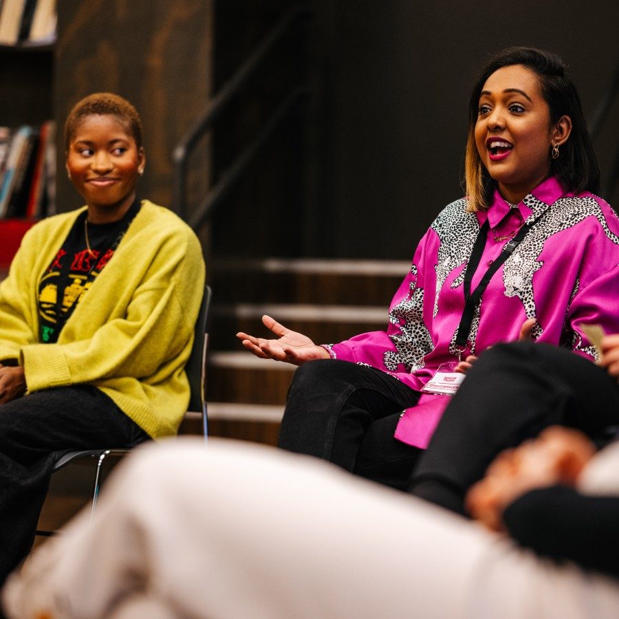 24-Theatreworks-PersonalImpactYellowPink-MatthewKaltenborn-2000x1000 Two women sitting on chairs in a rehearsal room in a circle with other people. The woman on the right is speaking and seems to be explaining something to the group. The other woman looks at her and listens.