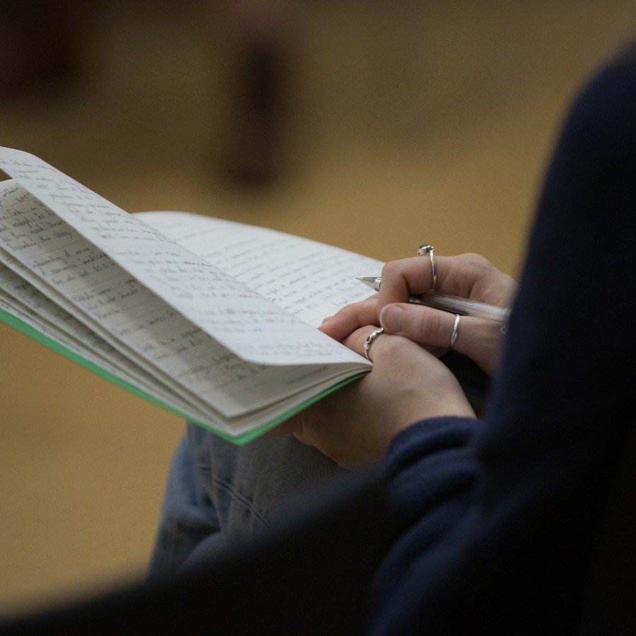 24-YoungProducers-Bookinhand-photographercredit-2000x1000 Person writing in an open notebook with handwritten text, holding a pen in their right hand. The foreground focuses on the writing, while the background is blurred, making it unclear.