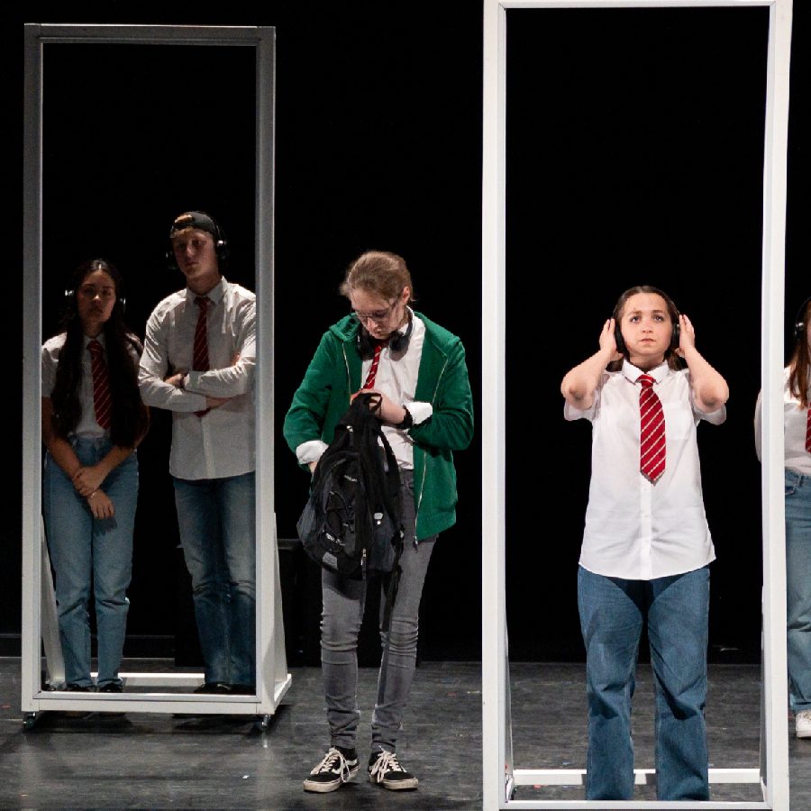 280624_Replica_Connections_JamieLee A stage performance depicting several people, each framed individually by rectangular door-like structures. The people are dressed casually, with some wearing white shirts and red ties. The scene is theatrical, set against a dark backdrop with minimal props.