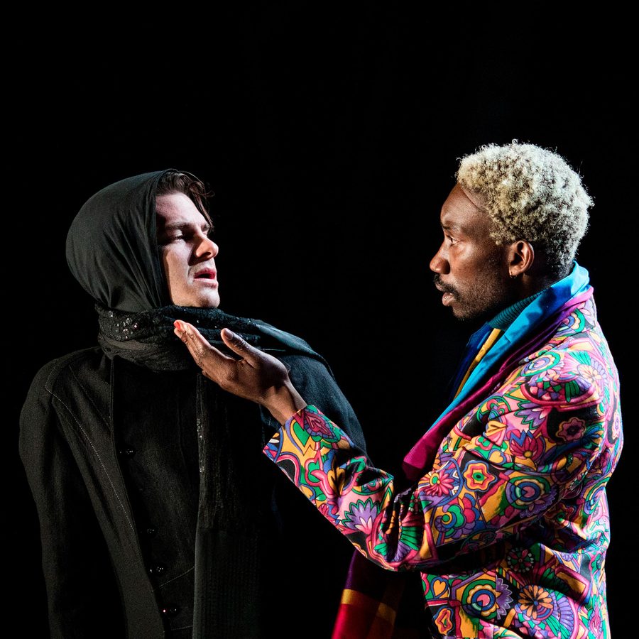 Angels-In-America-Part-2-2000x1000 Two actors in a powerful scene: one is dressed in black with a hood, looking serious, while the other, with blond hair, wears a vibrant, colorful jacket, gesturing expressively against a dark background.