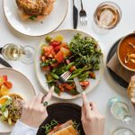 A table with lunch dishes of salads, savoury tarts, salmon beigel and soup. and glasses of wine.