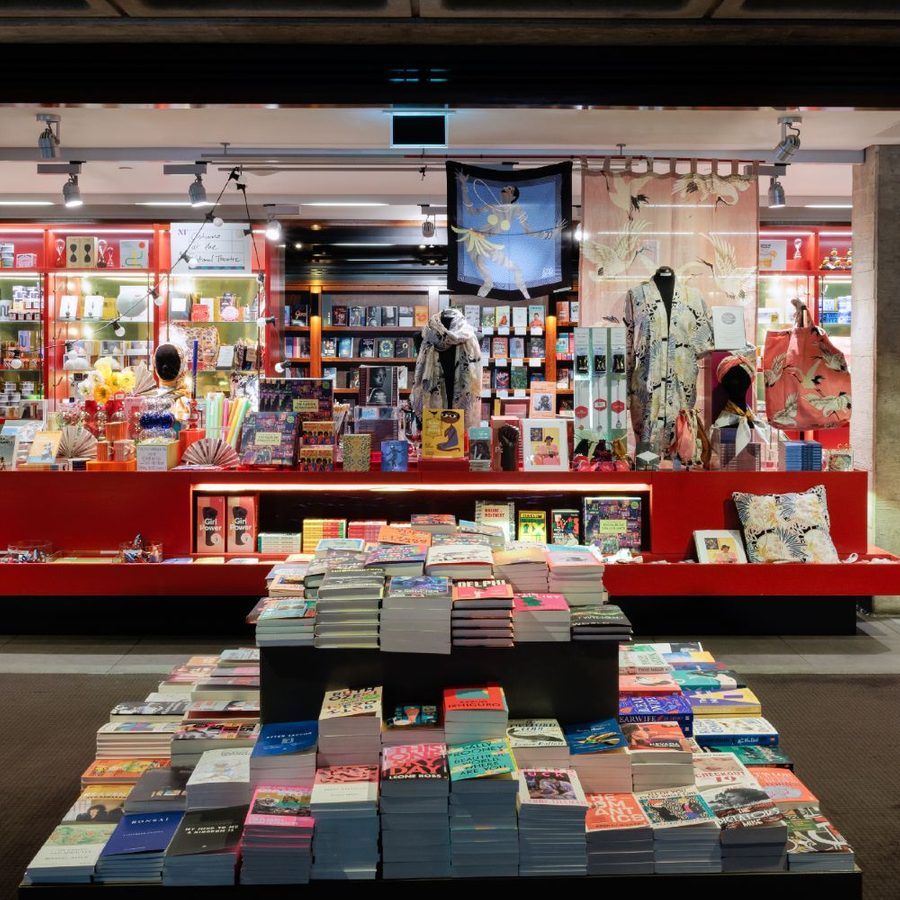Bookshop-masthead-desktop-v2024 A brightly lit bookstore with numerous books on tables and shelves. The central display features colorful children's books and decorative items. Shelves on the sides are filled with books of various genres, and a large open space leads to the back of the store.