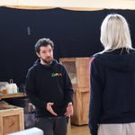 A woman stands and wears a black hoodie. A man speaks to her in a black hoodie.