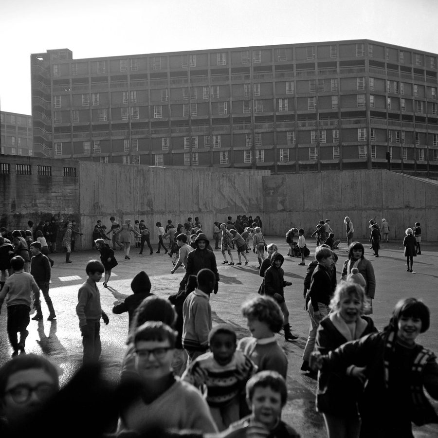 Children in the playground of Park Hill Junior School, 1969 About one hundred children play in a large concrete area. There is a brutalist building in the background. Black and white photograph.