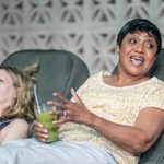 Two women sitting on green lounge chairs, one looking animatedly to the side holding a green juice.