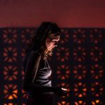 A woman looking down at her phone in the dar, she is lit by the light of her phone. Behind her is a patterned wall with an orange glow.