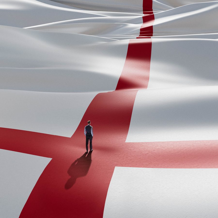 Dear-England-National Theatre-2000x1000-Masthead A lone figure stands, his back to us, wearing a waistcoat, shirt and trousers, in the centre of the red cross of the England flag that appears as a landscape rolling off into the distance.