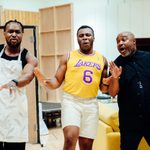 Three men in a rehearsal room, rehearsing a scene. Behind them are various pieces of furniture as part of a makeshift set. They are all expressive and animated.