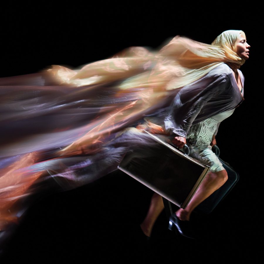 Kin-National-Theatre A woman runs, carrying a black suitcase with metal trim, her coat and headscarf billowing and blurred, behind her.