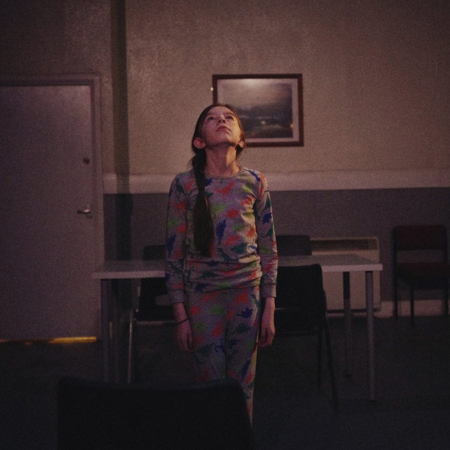 LOVE-production-image-masthead A young girl stands straight, in a dimly lit functional room, with a doorway off to the right open onto an illuminated space.