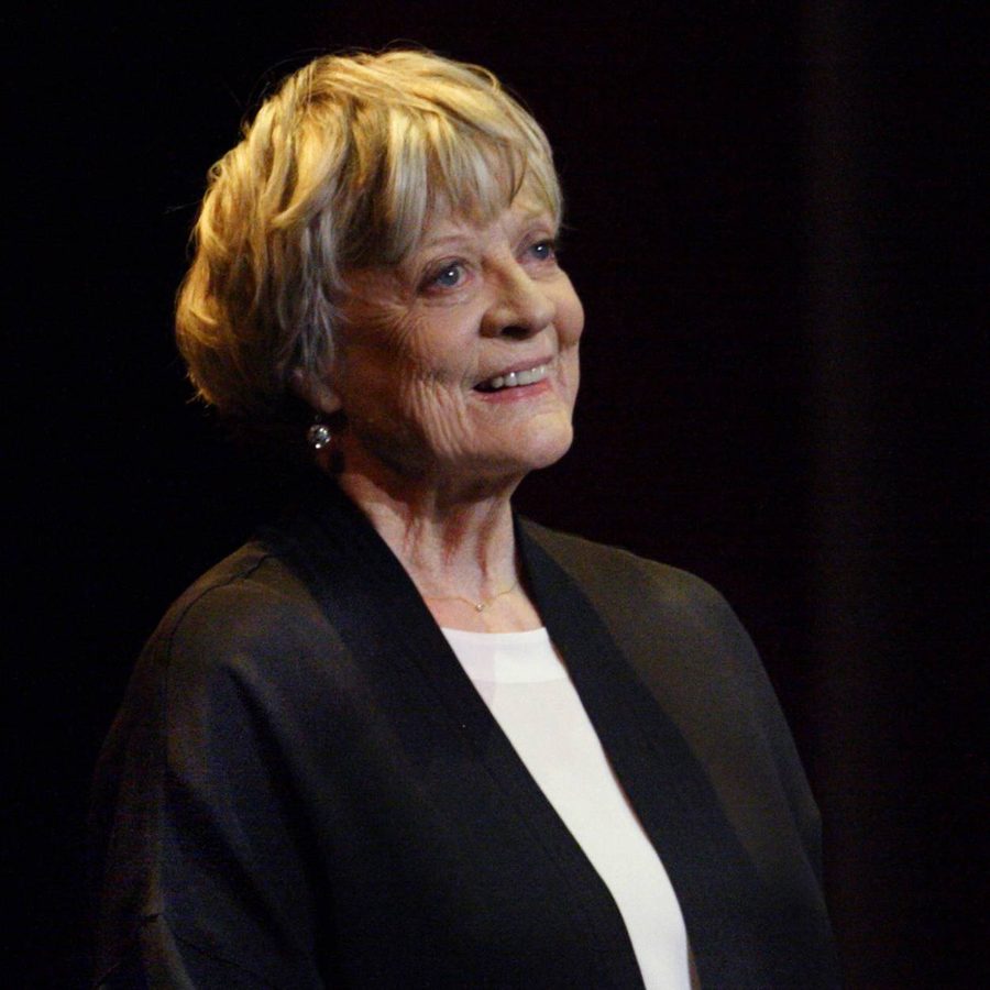 Maggie_Website_2000x1250_All_0000s_0003_50-Years-on-Stage-photo-Catherine-Ashmore-aspect-ratio-2000-1000 An older woman with short, tousled hair smiles while looking slightly to the side. She wears a dark jacket over a white top. The background is dark, emphasizing her presence.