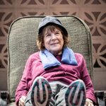 A woman smiling whilst lying down on a green lounge chair. She is wearing a purple longsleeve top, a blue neck pillow, blue hat and blue and white trainers.