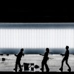 Five actors hold the same dramatic walking pose on a high-contrast stage with a bright horizontal light panel in the background. Multiple large rocks are strewn about the stage, and at the centre a wooden frame similar to a human body is laying on the ground.