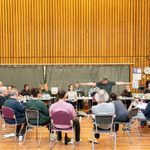 Twelve actors in a rehearsal room sat on chairs in a circle around their director. The director has his arms outstretched in demonstration. Behind them, eight people are sat at tables at the side of the room, with notes and laptops in front of them.