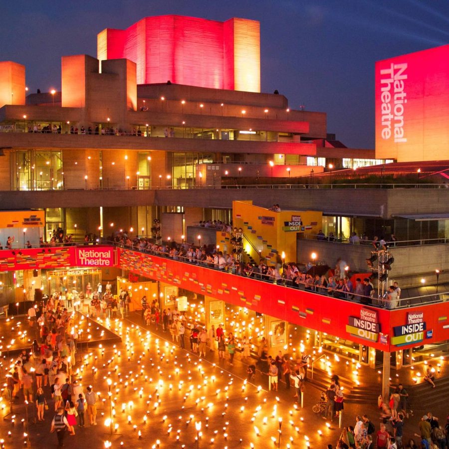 National-Theatre-Inside-Out-festival-with-flames A brightly lit National Theatre at night, featuring warm orange and red hues. The exterior is decorated with numerous lights, and people are gathered outside. The building's modern architecture is highlighted under the evening sky.