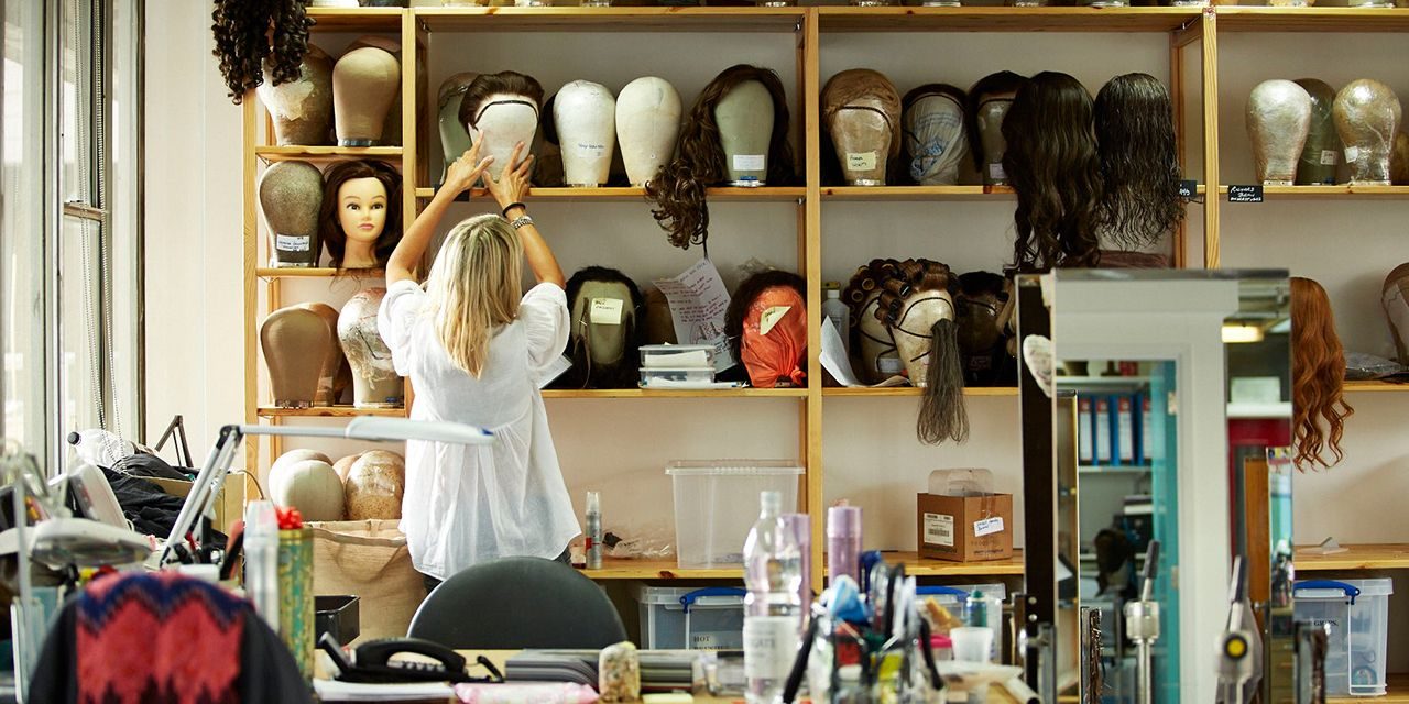 nt-wigs-hair-and-make-up-1280x640 A person arranges wigs on a shelf in a room filled with various wig mannequins, styling heads, and hairpieces. The workspace is filled with styling tools, bottles, and hair products, creating a busy, organized environment.