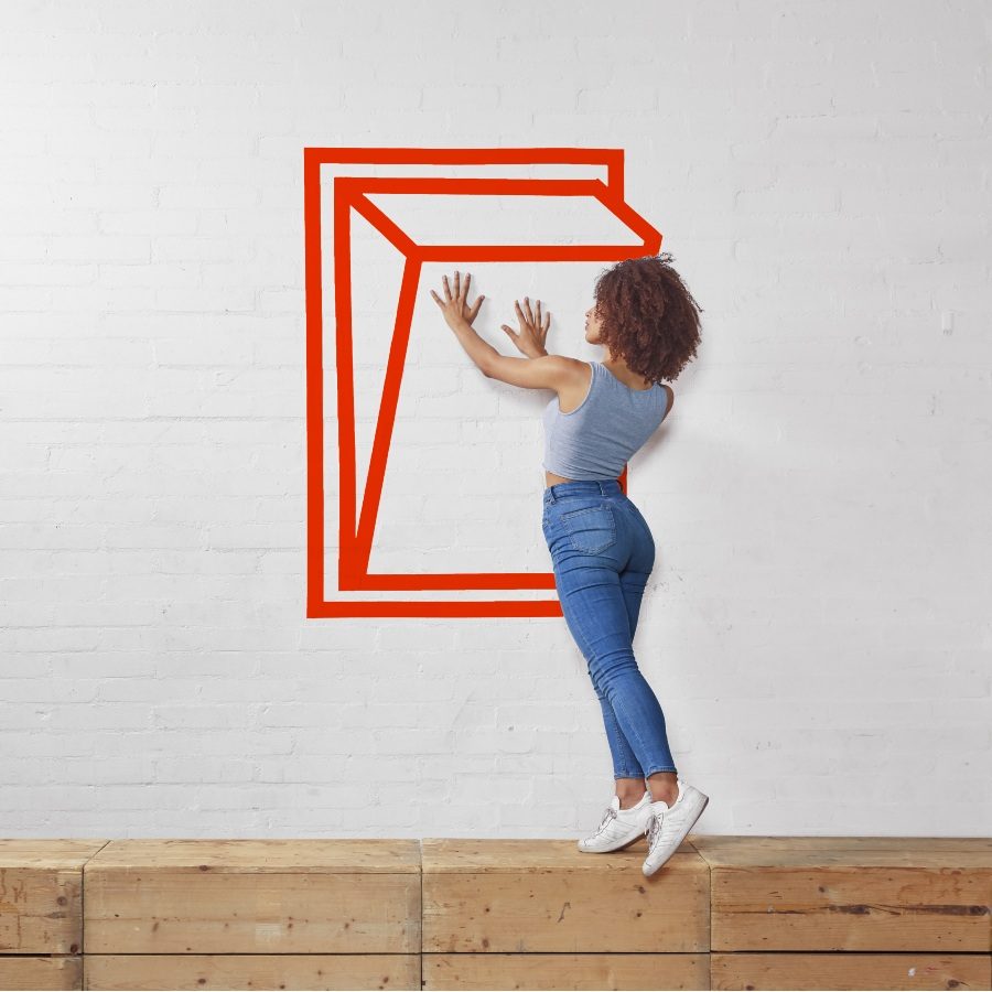 NTGDS_NF_TheatreWorks_Web Header__2000x1000px_4 reduced Three people interact playfully with red geometric tape designs on a wall. Each person appears to be inside or interacting with a 3D cube while standing on wooden crates. The cubes create an optical illusion effect.