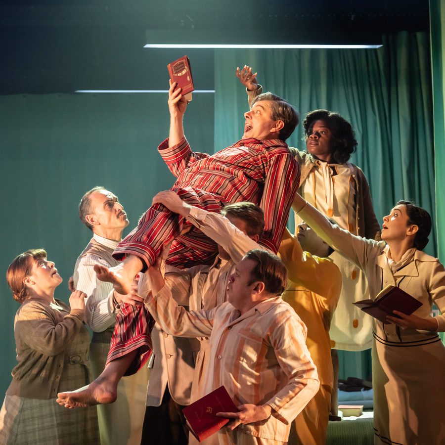Nye-cast-National-Theatre-Johan-Persson-2-scaled-aspect-ratio-2000-1000 Michael Sheen lifted into the air by other cast members, wearing red striped pyjamas.