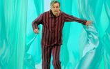 Nye-Michael-Sheen-National-Theatre-2025-mobile A man wearing red and black striped pajamas and plaid slippers is moving through flowing, translucent teal curtains. He extends his left arm forward, to part one of the curtains. The background consists of soft, undulating teal curtain layers, creating an abstract, dreamy environment.
