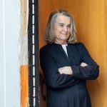 A woman with long grey hair, dressed in a black suit, stands with crossed arms leaning against a brown door, set in a frame with black vertical stripes, surrounded by light grey walls.