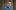 rufus-norris-2000x1000-masthead-photo-by-cameron-slater-photography-01-aspect-ratio-160-100 A man with short brown hair wearing a blue button-up shirt stands in front of a textured, abstract background with vertical lines. He has a neutral facial expression and is looking directly at the camera.