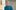 rufus-norris-2000x2000-masthead-photo-by-cameron-slater-photography-aspect-ratio-160-100 A man with gray hair, wearing a blue button-up shirt, is smiling while sitting indoors. The background is slightly blurred and appears to be a room with beige walls and a closed door.