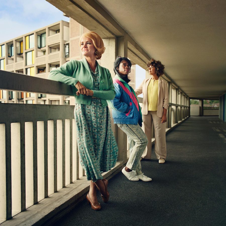 Standing-at-the-Skys-Edge-2000x1000 Three women, of mixed age and race, leaning against railings on a walkway in a block of flats.