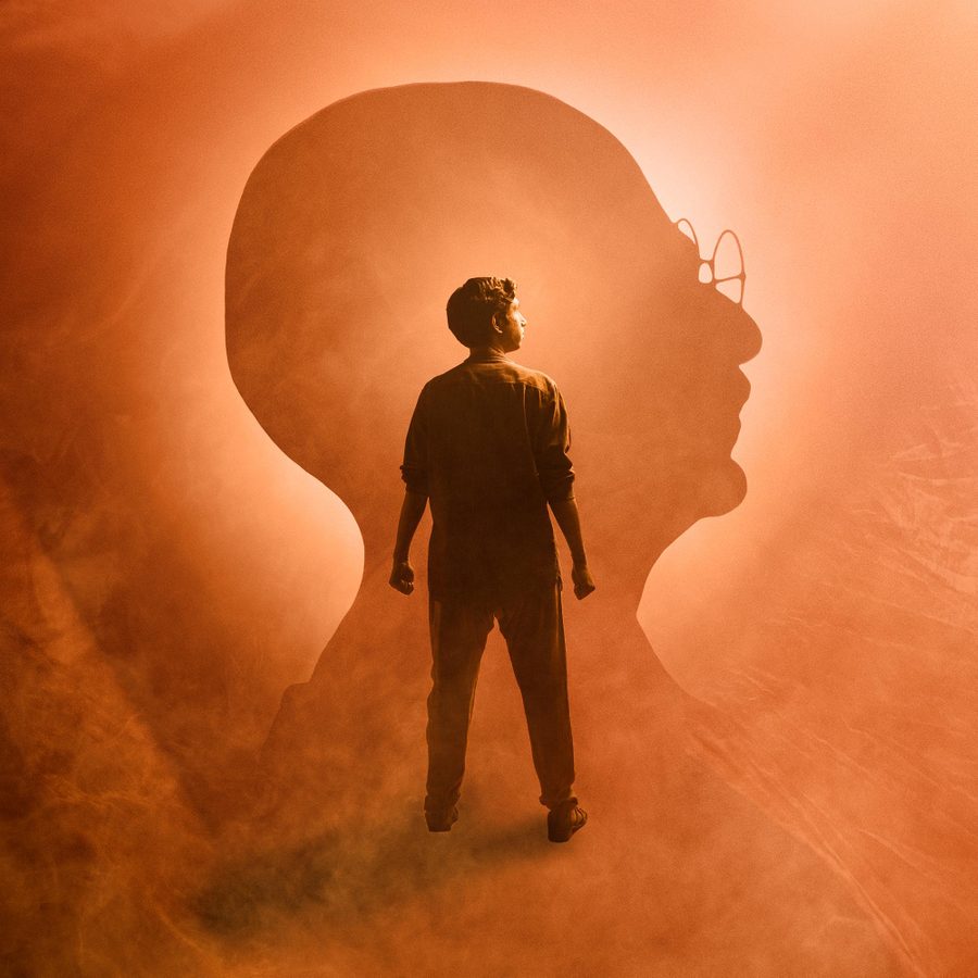 The-Father-and-the-Assassin-2023-National-Theatre-2000x1000 A photo-montage of a figure of a young man standing, arms by his side, hands balled into fists, and behind him a large profile of Gandhi, with both heads looking off to the right.