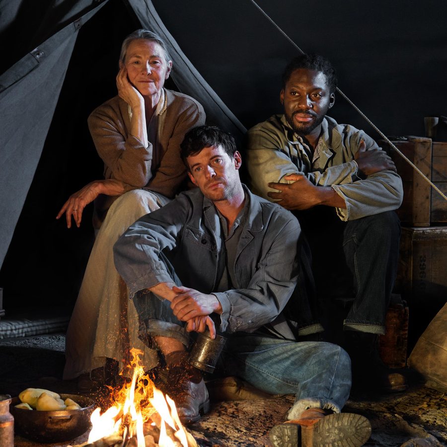 The-Grapes-of-Wrath-National-Theatre-artwork-v2-masthead A woman and two men sit together in front of the open flap of a canvas tent. Packing boxes and canvas bags are either side of them and an open fore is before them.