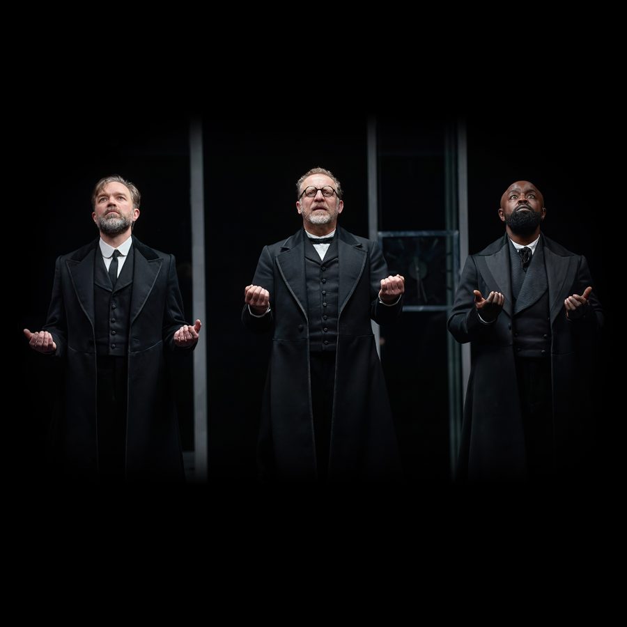 The-Lehman-Trilogy-Gillian-Lynne-Theatre-Production-Image-2000x1000 Two white men and a black man wearing black 3-piece suits stand with their hands raised in front of them, looking upwards with sombre expressions.