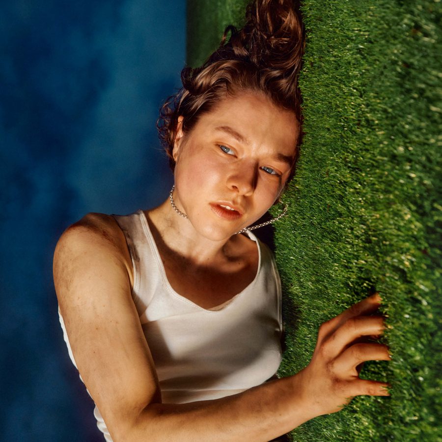 The-Other-Place-National-Theatre-image-v2-2000x1000 A person with short brown hair wearing a white tank top lies on their side on a grassy surface looking slightly upward. The image is taken from an angle that makes it appear as if the person is standing against a vertical grass wall, with a blue sky background.