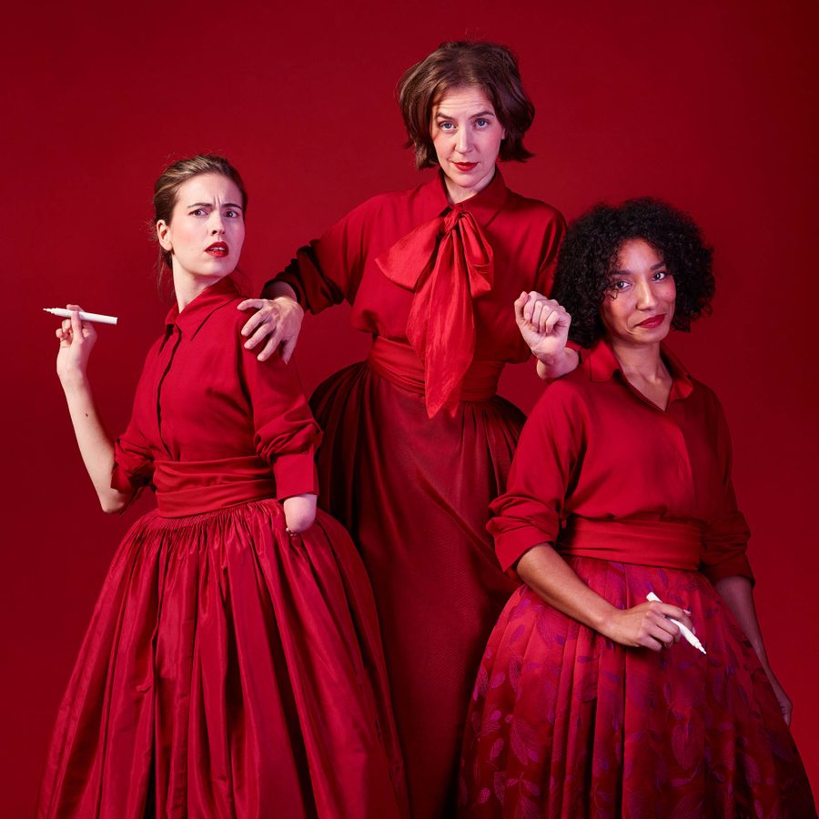 Underdog-National-Theatre-Northern-Stage-2000x1000-1b Three women stand together, all facing to camera, the middle one, slightly higher than the others, seems to be trying to push the others, who each hold a pen, aside. All wear red shirts and skirts and the middle one has a large red bow tied at her collar.