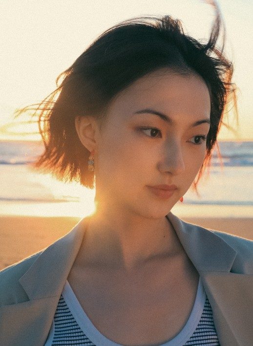 Yimei-Zhao-headshot-HanxiaoLuo-518x708 A person with black hair in a bob haircut, standing against a sunset on the beach